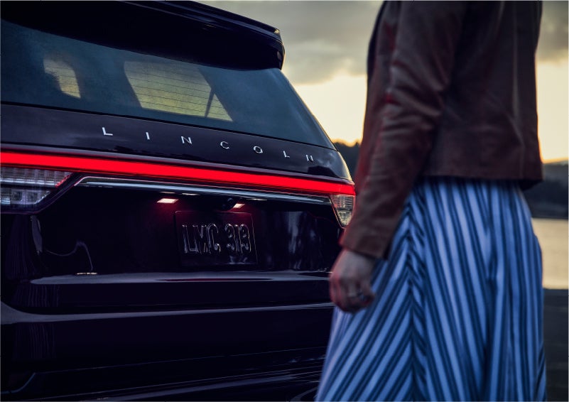A person is shown near the rear of a 2023 Lincoln Aviator® SUV as the Lincoln Embrace illuminates the rear lights | Eau Claire Lincoln in Eau Claire WI