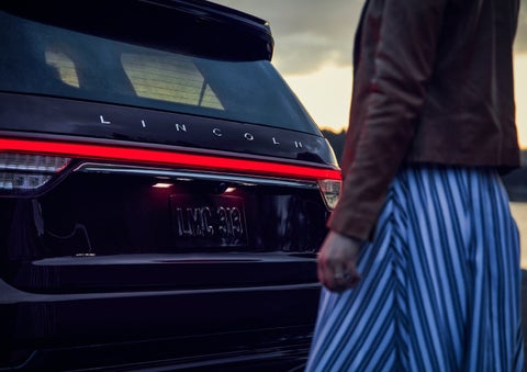A person is shown near the rear of a 2024 Lincoln Aviator® SUV as the Lincoln Embrace illuminates the rear lights | Eau Claire Lincoln in Eau Claire WI