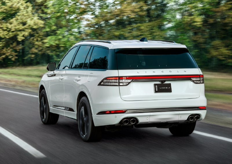 A white 2026 Lincoln Aviator® SUV is being driven away down a road lined with colorful trees. | Eau Claire Lincoln in Eau Claire WI