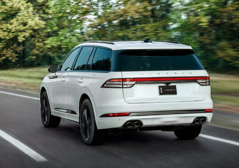 A white 2026 Lincoln Aviator® SUV is being driven away down a road lined with colorful trees. | Eau Claire Lincoln in Eau Claire WI